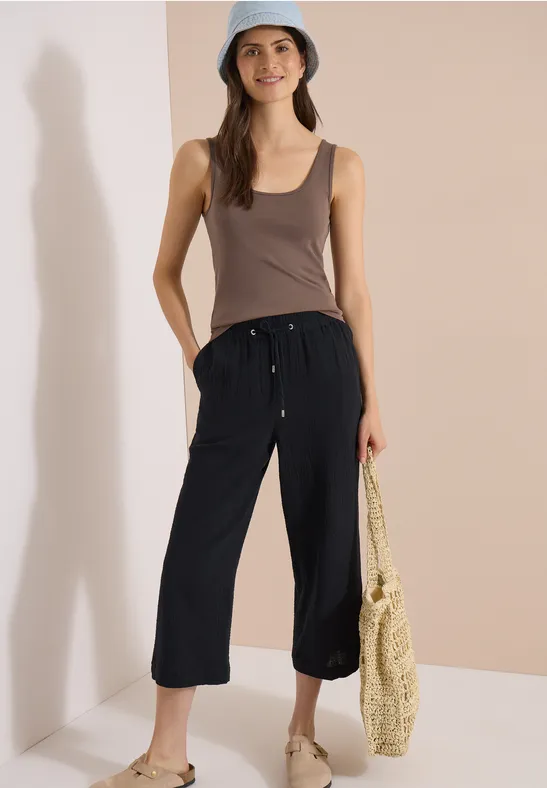 Frau in schwarzer 3/4-Hose, braunem Top, Strohtasche und Bucket Hat vor schlichtem Hintergrund.