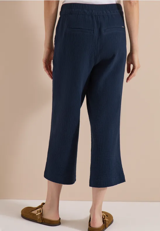 Achteraanzicht van persoon in wijde, donkerblauwe enkellange broek met zakken en bruine mocassins voor beige achtergrond.