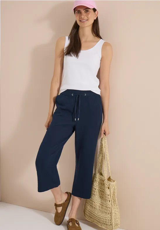 Een vrouw draagt een wit topje, donkerblauwe culottes, sandalen en houdt een strooien tas vast.