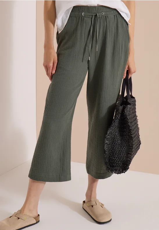 Frau trägt olivgrüne weite Culotte-Hose mit Tunnelzug, beigen Mules und schwarze Netz-Tasche
