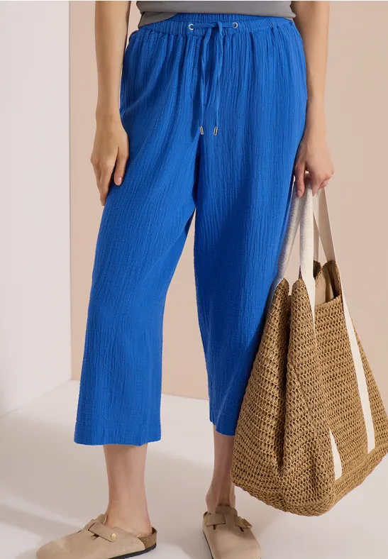 Eine Person trägt knöchellange, weite, blaue Hose mit Kordelzug, flache Sandalen und hält eine Strohtasche.