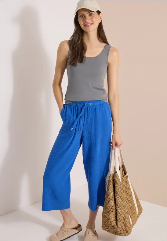Frau in grauem Tanktop und blauer Culotte, Sandalen und geflochtener Tasche vor hellem Hintergrund.