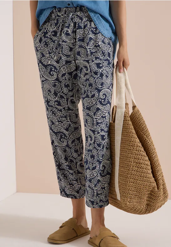 Femme en pantalon bleu à motif cachemire, lien à la taille, sandales beiges et sac en osier.