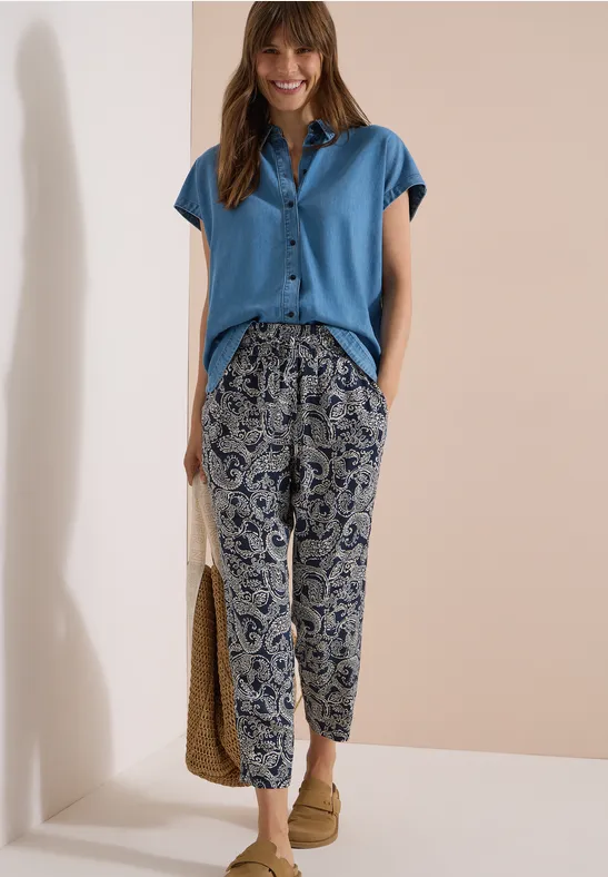 Femme en chemisier bleu à manches courtes, pantalon large à motif, mocassins clairs et sac en paille.