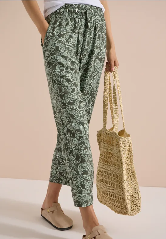 Eine Person trägt grüne, locker sitzende Paisley-Hose, beige Slipper und hält eine geflochtene Strohtasche.