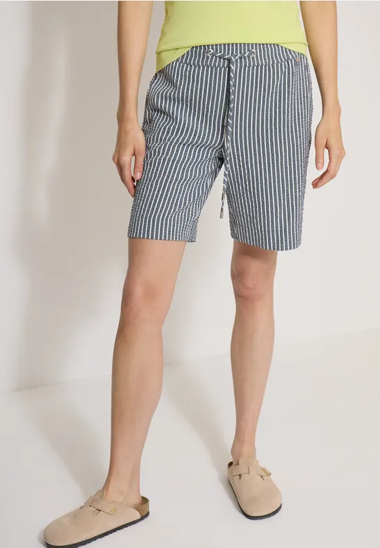 Frau trägt kniehohe, blau-weiß gestreifte Shorts mit Knopfleiste und beige Hausschuhe.