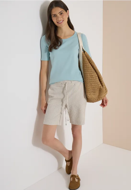 Eine Frau trägt ein hellblaues T-Shirt, beige Shorts, braune Schuhe und eine Strohtasche und lehnt an einer Wand.