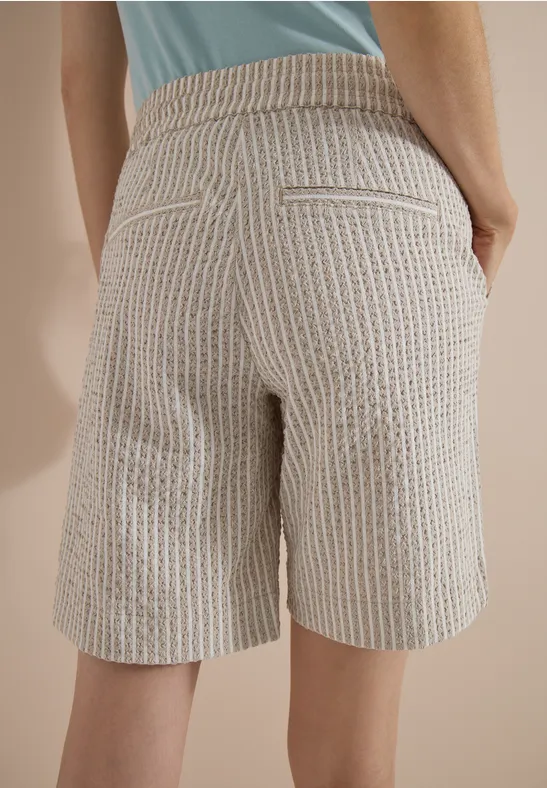 Rückansicht einer Person in beige-weiß gestreiften Bermudashorts mit Taschen und hellblauem Oberteil vor beige Hintergrund.