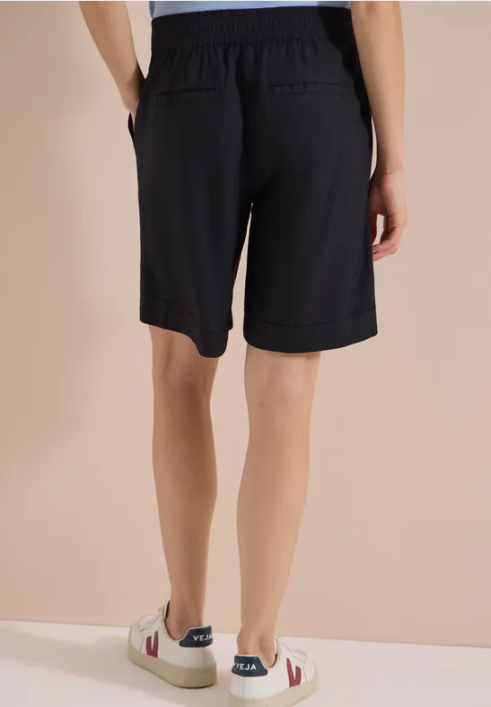 Achteraanzicht van een persoon met zwarte short en witte sneakers voor een neutrale achtergrond.
