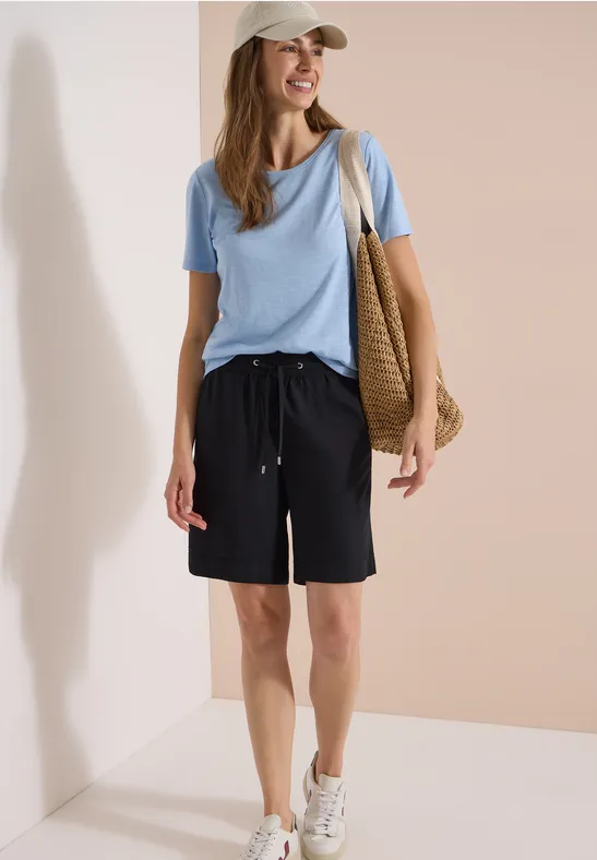 Vrouw in blauw t-shirt, zwarte bermudashorts, witte sneakers, pet en rieten tas, tegen neutrale achtergrond.