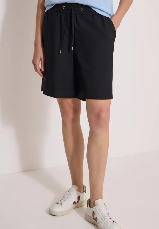 Model draagt zwarte, knielange short met koord en witte sneakers.