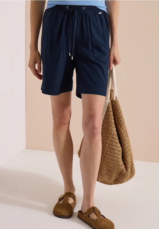 Frau trägt marineblaue Shorts mit Kordelzug und braunen Sandalen, hält eine geflochtene Tasche.