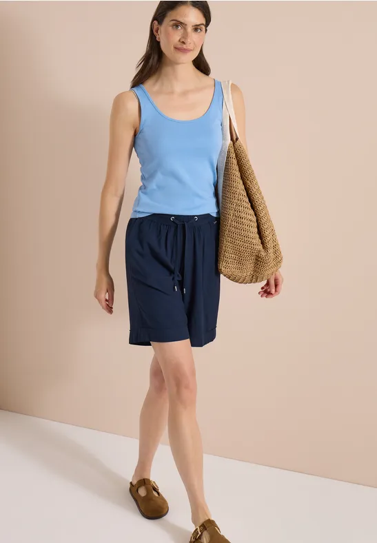 Frau in blauem Tanktop, dunkelblauen Shorts und braunen Sandalen vor beigem Hintergrund mit großer Gewebetasche.