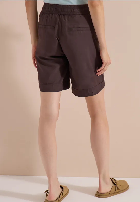 Von hinten gezeigte Person in dunkelbraunen Shorts und grünen Slippern vor beigem Hintergrund.