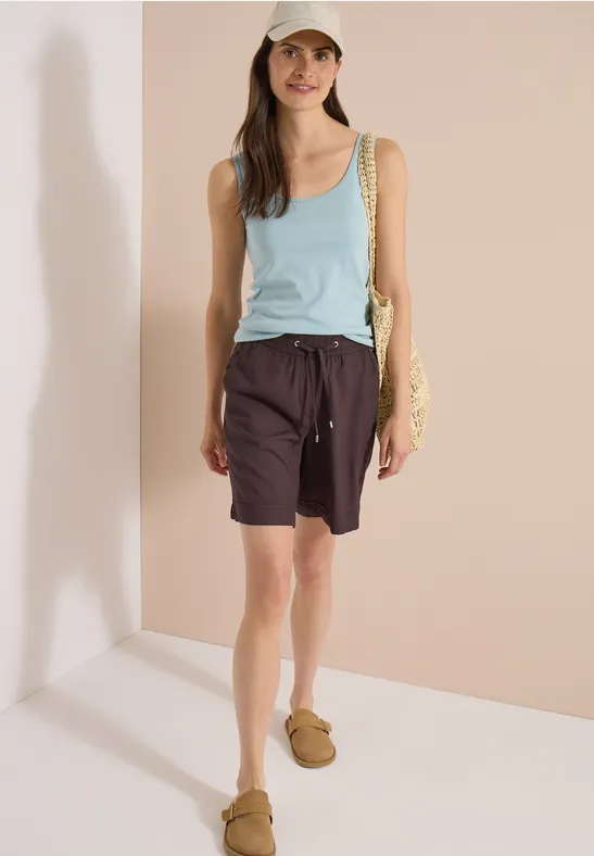 Frau in türkisfarbenem Tanktop, braunen Shorts mit Kordelzug und braunen Sandalen vor hellem Hintergrund.