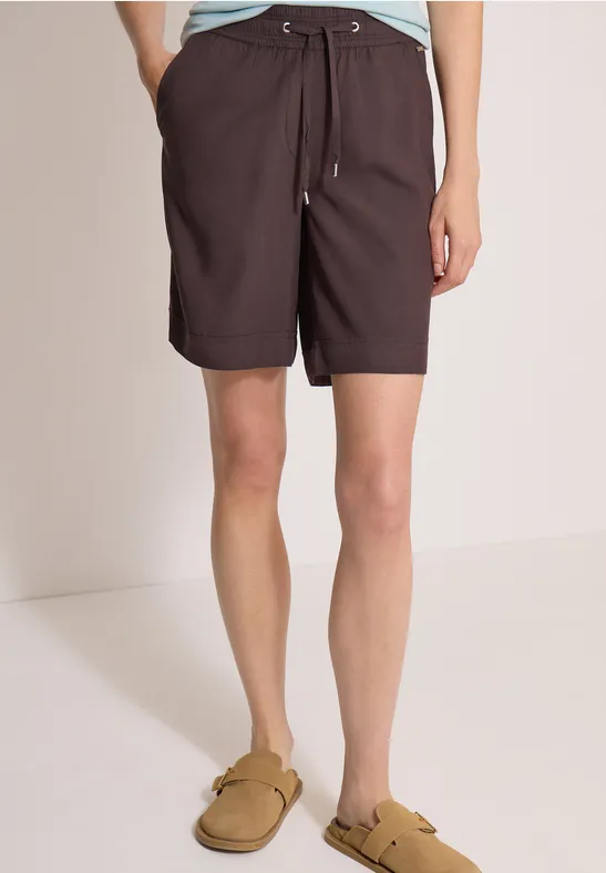 Eine Person trägt dunkelbraune Shorts mit Kordelzug und beigefarbene Slip-on-Sandalen.