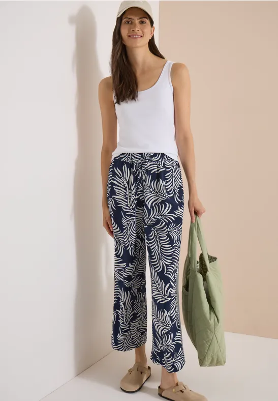 Vrouw met wit hemdje en wijde blauw-witte broek met palmbladprint, groene tas en beige schoenen.