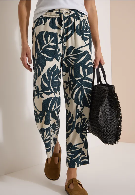 Weite, helle Hose mit dunkelgrünem Blattmuster, braune Loafers und schwarze, geflochtene Tasche.