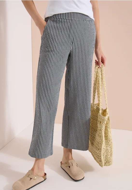 Persoon in wit shirt, gestreepte, wijdvallende 7/8-broek, beige loafers en een gevlochten rieten tas.