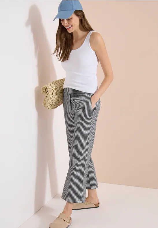 Vrouw in witte top, grijze geruite broek, blauwe pet en stro tas leunend tegen een muur.