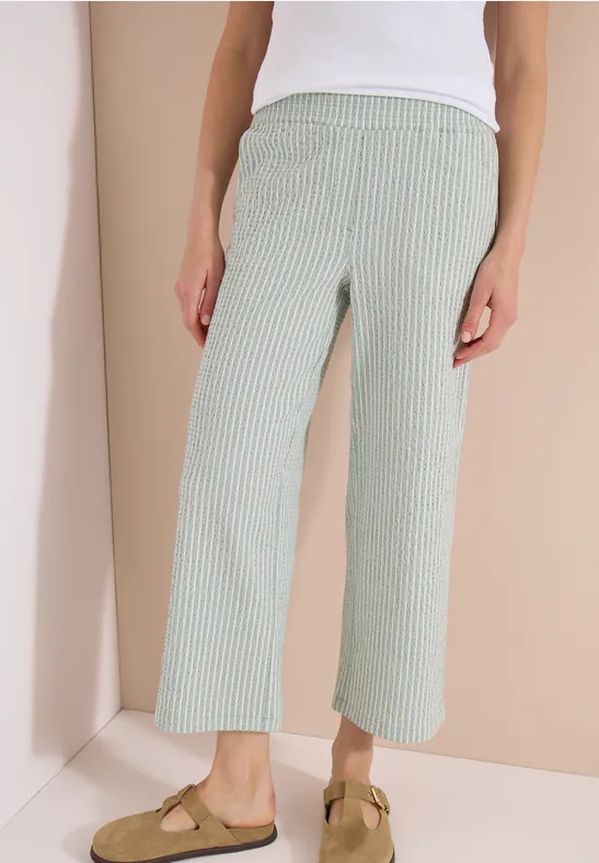Vrouw in wit topje, groen-wit gestreepte culotte, beige mocassins met kwastjes.