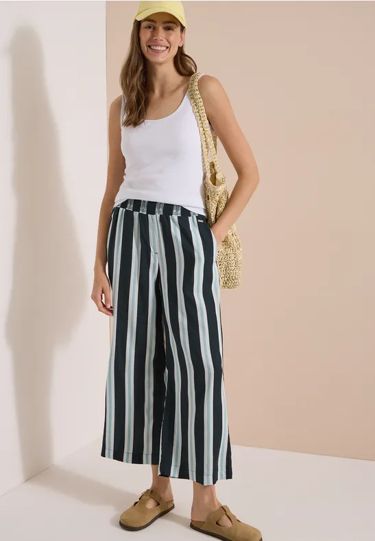 Vrouw in witte tanktop, brede gestreepte broek, strooien tas en sandalen voor een lichte achtergrond.