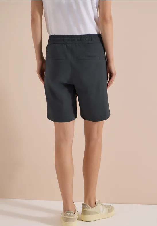 Achteraanzicht van een persoon in een wit topje, zwarte short en beige sneakers voor een lichte achtergrond.