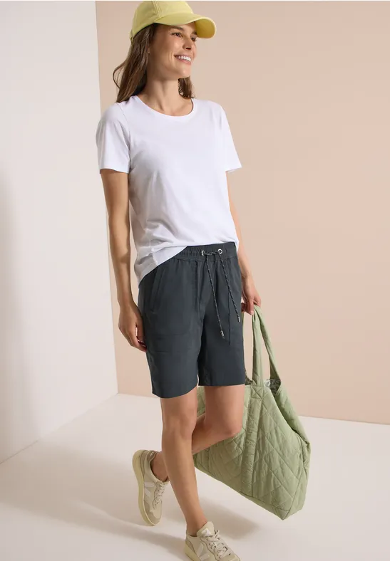 Vrouw in wit t-shirt, grijze short en beige sandalen met een groene tas.