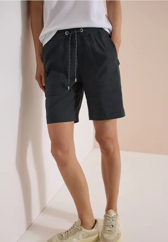 Persoon in zwarte shorts met trekkoord, witte top en lichte sneakers tegen een minimalistische achtergrond.
