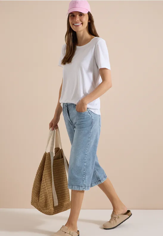 Frau in weißem T-Shirt und Jeans-Culottes mit Strohtasche und Slippern.