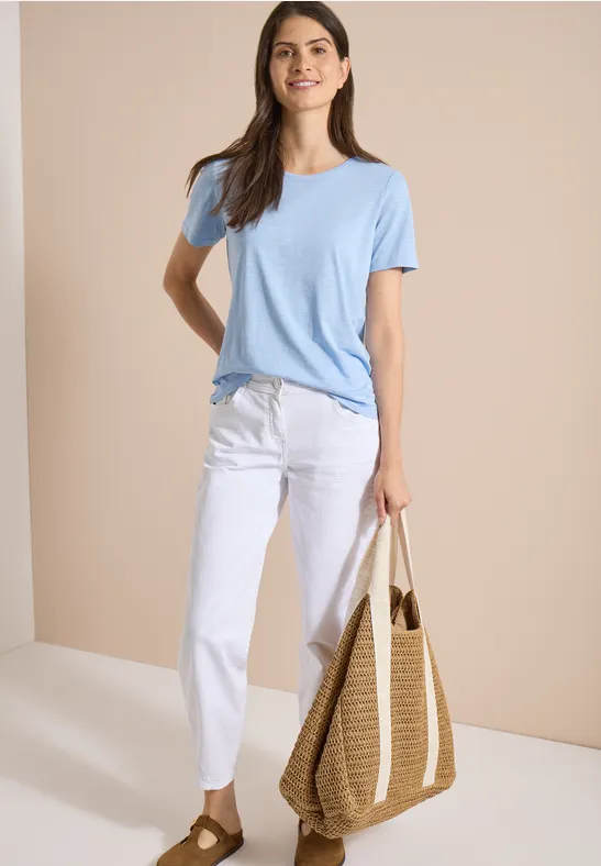 Vrouw met lichtblauw t-shirt, witte broek en rieten tas, voor een beige achtergrond.