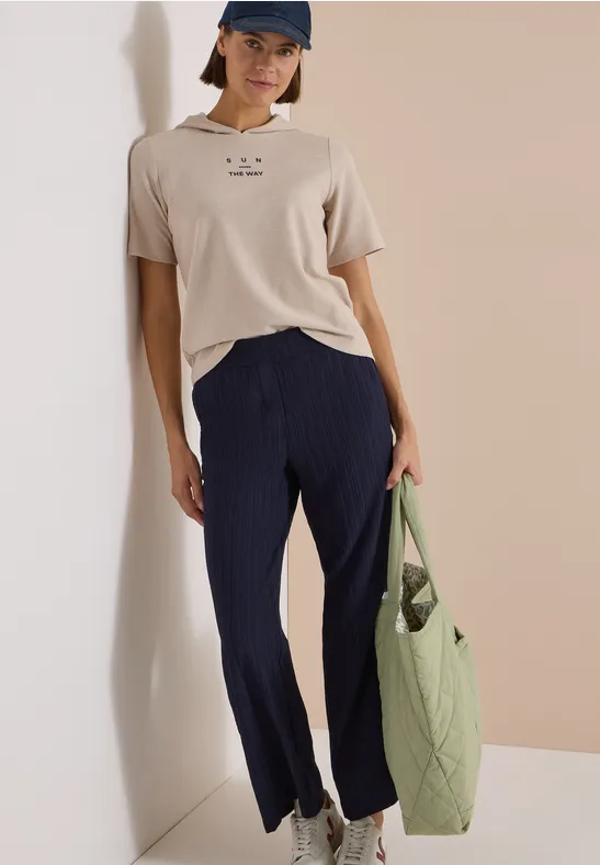 Frau trägt beige T-Shirt, dunkelblaue Hose, blaue Kappe und hält olivgrüne Stepp-Tasche.