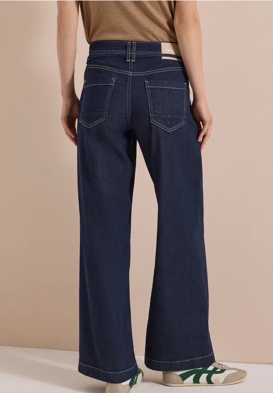 Frau von hinten in hoher Taille, weiten dunkelblauen Jeans und weißen Sneakers mit grünen Details.