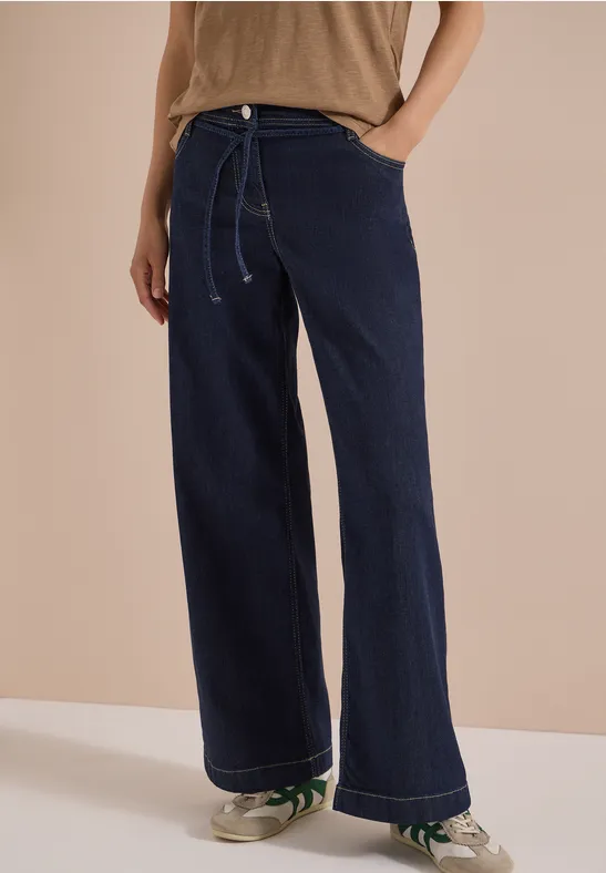 Frau trägt weite, dunkelblaue Jeans mit Kordelzug und Sneakers vor hellem Hintergrund.
