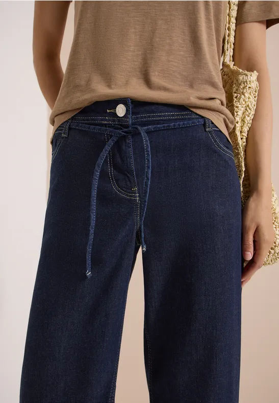 Person trägt hoch geschnittene, dunkelblaue Jeans mit Bindegürtel, kombiniert mit braunem Oberteil und Basttasche.