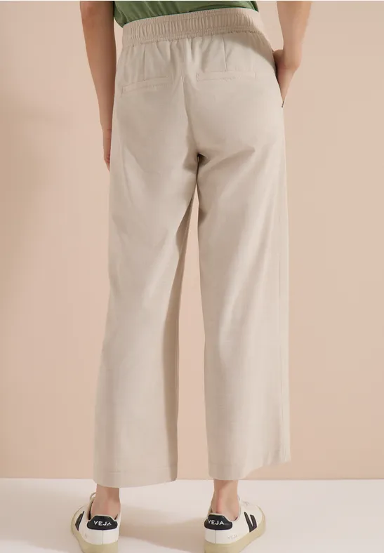 Achteraanzicht van iemand in beige, wijde broek met elastische taille en zwarte espadrilles.