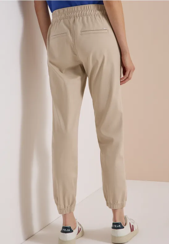 Vue de dos d'une personne portant un pantalon jogger beige avec taille et chevilles élastiquées près d'un mur clair.