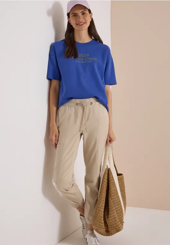 Femme souriante en T-shirt bleu, pantalon beige et grand sac en paille contre un mur clair.