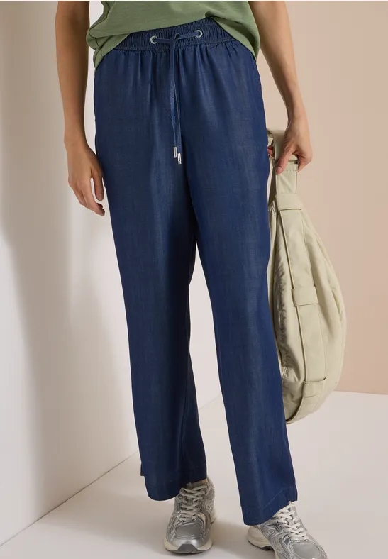 Person trägt weite, dunkelblaue Stoffhose mit Tunnelzug, silberne Sneaker und eine leichte Umhängetasche.