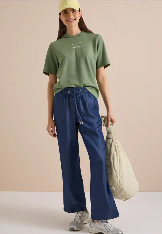 Junge Frau in grünem T-Shirt, weitem blauen Hosen und Cap, lässig stehend mit großer Tasche.