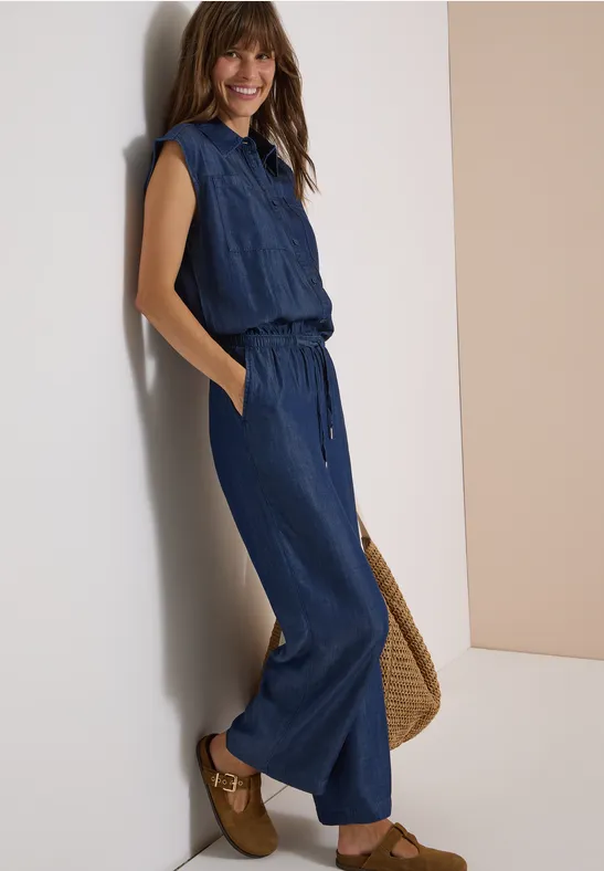 Eine Frau lehnt an einer Wand, trägt einen blauen Jeans-Jumpsuit, Basttasche und braune Loafers.