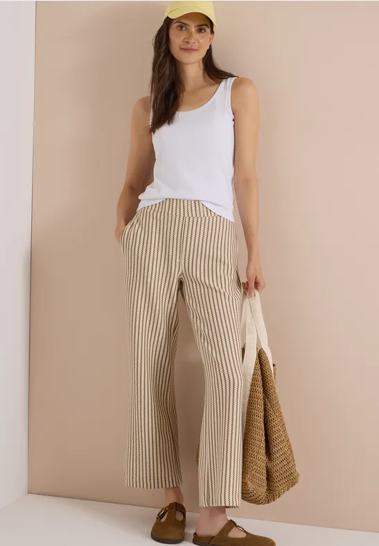 Frau trägt weißes Tanktop, beige-weiß gestreifte, weite Hose und hält eine geflochtene Tasche.
