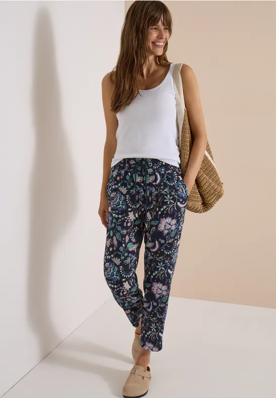 Frau in weißem Top, gemusterten Paisley-Hosen, Basttasche und beigen Slippern vor neutraler Wand