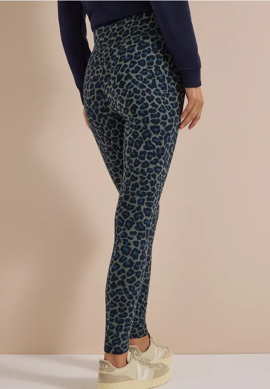 Een persoon draagt blauwe legging met donkerblauw luipaardpatroon en lichte sneakers voor een beige achtergrond.