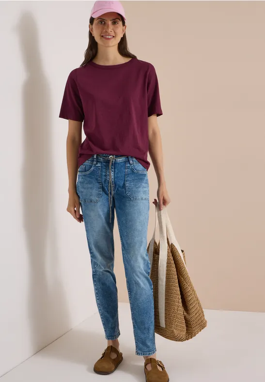 Eine Frau in rosa Cap, burgunderrotem T-Shirt, blauen Jeans und mit einer beigefarbigen Tasche vor hellem Hintergrund.