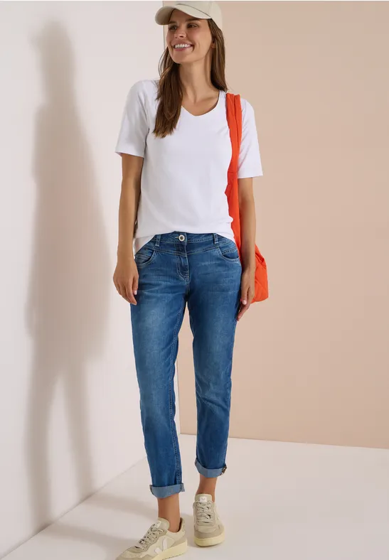 Frau in weißem T-Shirt, Jeans, Sneakers und Cap, mit orangem Schulterbeutel, steht lächelnd vor hellem Hintergrund.