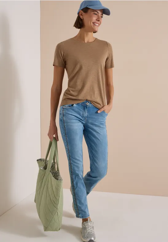 Frau in braunem T-Shirt, hellblauen Jeans, grauen Sneakers und blauer Kappe mit grünem Stoffbeutel vor beiger Wand.