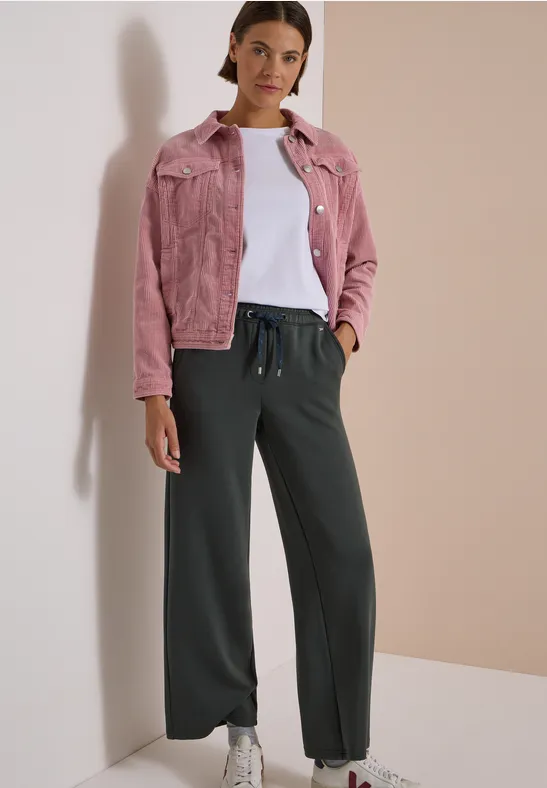 Frau vor neutralem Hintergrund mit rosa Jeansjacke, weißem Shirt, weiten schwarzen Hosen und weißen Sneakern.