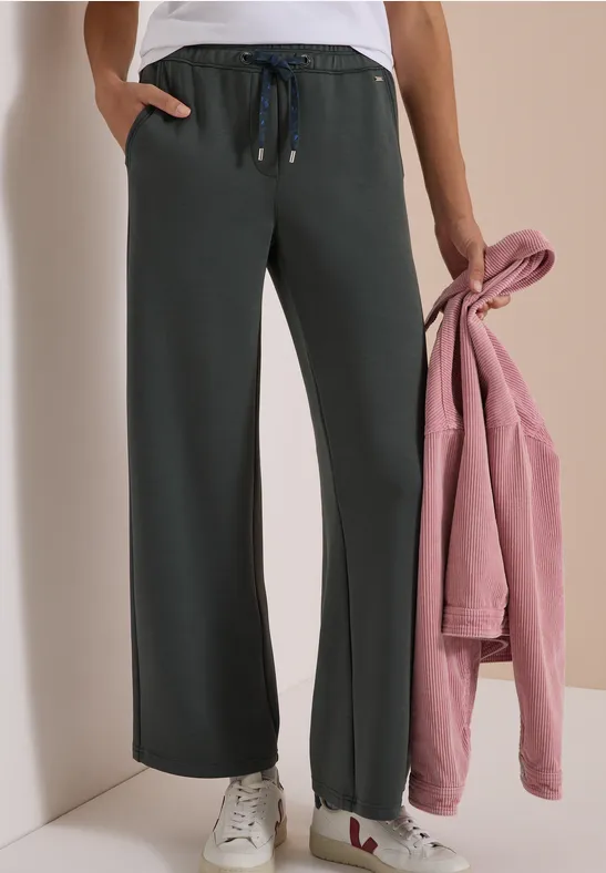 Dame trägt weite, dunkelgraue Jogginghose mit Tunnelzug, weiße Sneaker und hält ein rosa Sweatshirt.