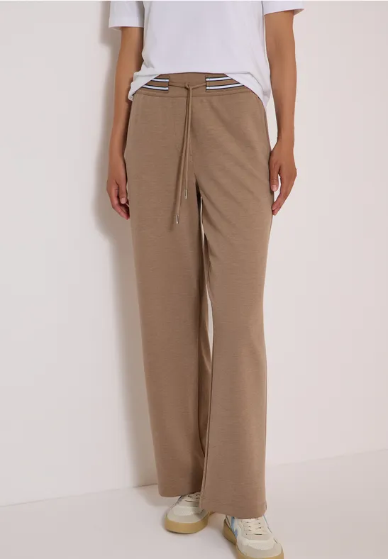 Weite hellbraune Cordhose mit Kordelzug, weiße Sneaker, Person vor weißem Hintergrund.